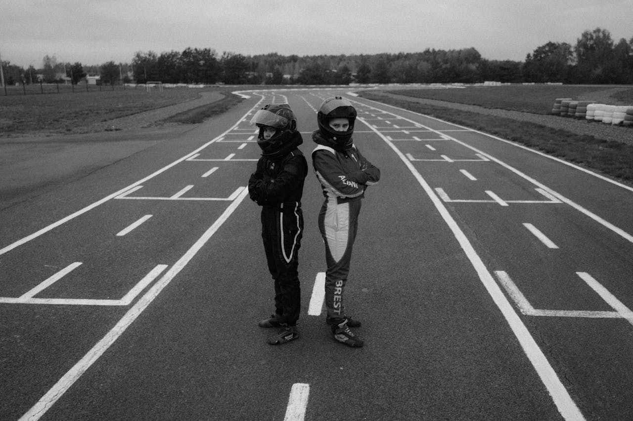 Two karting racers standing back-to-back on a racetrack in full gear, showcasing determination in monochrome.